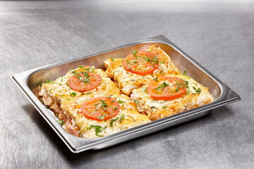 Layers of baked pasta fill a stainless steel tray, topped with fresh tomato slices and sprinkled with herbs, highlighting a savory dish prepared for lunch