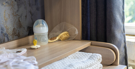 Baby clothes, feeding bottle, pacifier, and hairbrush are neatly arranged on a wooden changing table, indicating a parent getting ready for their newborn.