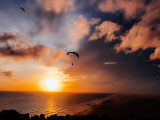 Silhouette of a crowd gathered on a high viewpoint, watching a sunset over the ocean. The dreamy glowing orange and blue sky reflects over the sea, creating a peaceful and majestic atmosphere.