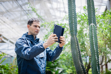 Fototapeta premium Young scientist in farm greenhouse is using tablet for agricultural research while focusing on cactus plants, dedicated research team effort in natural environment