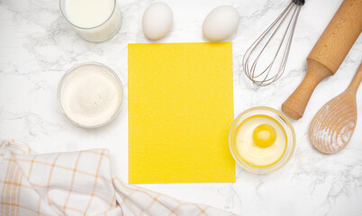 Bright yellow paper placed on a marble countertop surrounded by baking ingredients and utensils in...