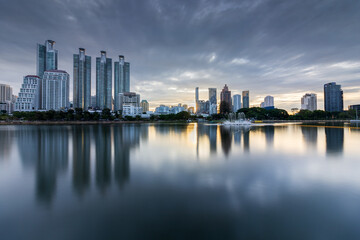 Fototapeta premium Peaceful sunrise view at Benjakitty Park in Bangkok, Thailand, with calm lake waters reflecting nearby skyscrapers