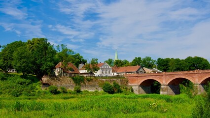 The historic brick bridge over the Venta River in Kuldīga, Latvia, with charming old town houses and a church spire surrounded by lush greenery under a bright summer sky.