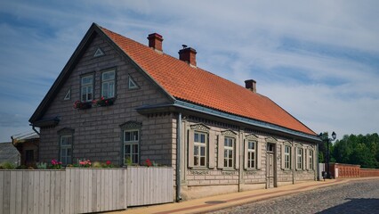 Traditional wooden house with decorative window frames and a red tiled roof, located on a cobblestone street with a wooden fence and flowers on a sunny day.