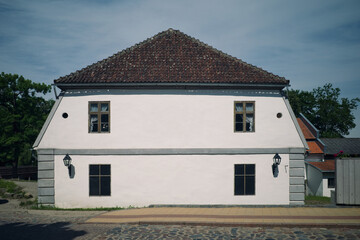Historic white building with a red tiled roof, symmetrical windows, and classic lanterns, located on a cobblestone street under a blue sky.