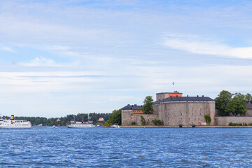 The historic stone Vaxholm Fortress stands surrounded by water and lush greenery, with a Swedish flag flying proudly atop its walls, rich heritage and scenic coastal location