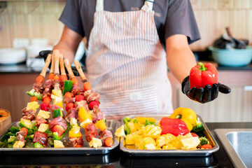 Chef holding a red bell pepper while preparing colorful meat and vegetable skewers, showcasing fresh ingredients and vibrant food preparation in a home kitchen setting.