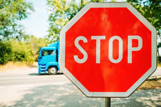 Bright red stop sign in sharp focus on a sunny day, with a blurred blue truck passing by in the background. Set against green trees and a quiet road.