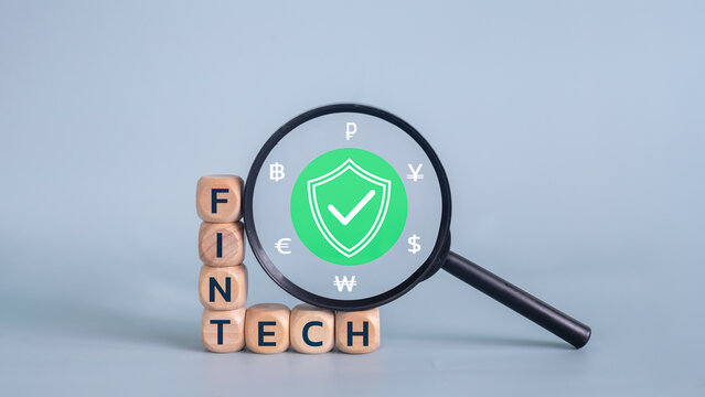 Wooden blocks spelling FINTECH with a magnifying glass, focusing on green security shield and global currency symbols. Concept of financial technology and secure digital payments.