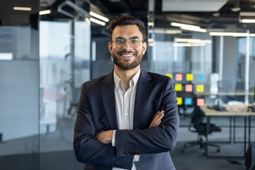 A confident businessman in a suit poses for a photo with a warm smile in a modern office setting.
