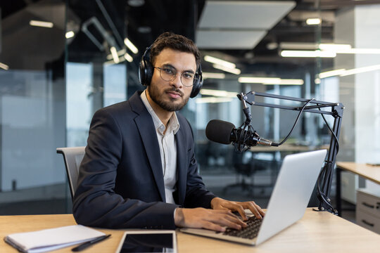 A businessman in a suit wearing headphones and glasses sits at a desk with a microphone, laptop, and tablet in an office setting. - Powered by Adobe