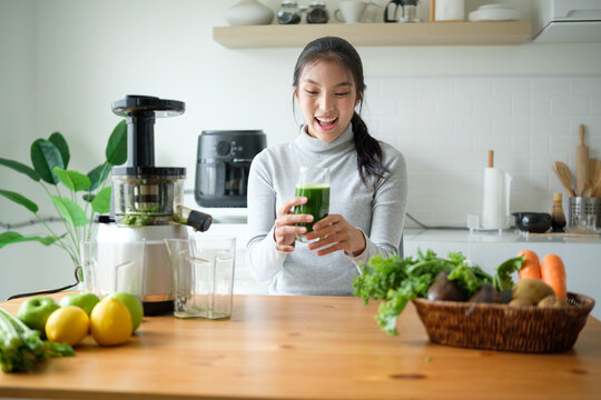 Happy woman holding a glass of fresh green juice in a bright modern kitchen with juicer and vegetables on the counter. - Powered by Adobe
