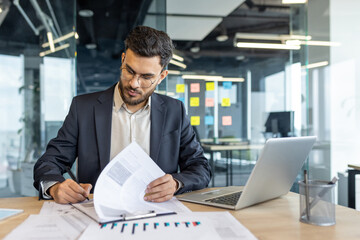 A businessman is working in his modern office, reviewing documents and using his laptop. Focused and professional, he's dressed in a suit.