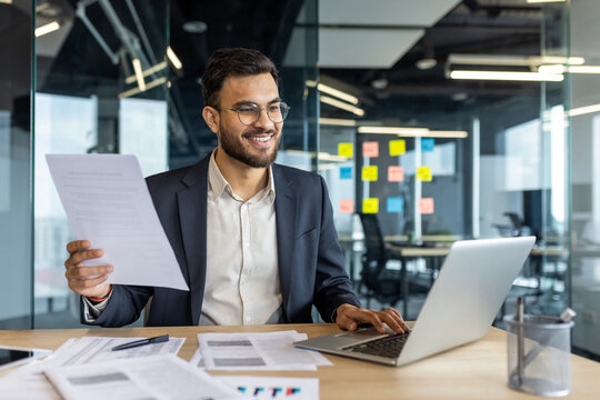 A smiling businessman reviews documents while working on a laptop in a modern office environment, focused on productivity.