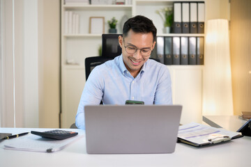 Smiling businessman using smartphone while working on a laptop at a modern office desk.
