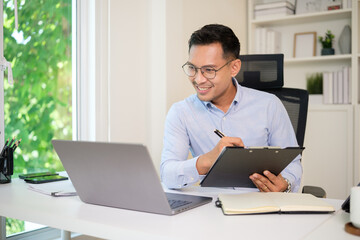 Businessman smiling while working on a laptop and writing notes on a clipboard in a modern office.