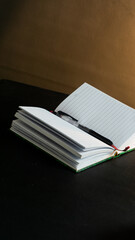 Portrait side view of notebook and pen on book under warm light, placed on black table with black background, minimal and cozy dark theme setup