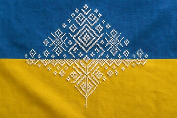 A digitally composited Ukrainian flag where the azure half transitions into traditional vyshyvanka embroidery patterns (white geometric motifs), while the yellow half remains smooth. Overhead flat lay