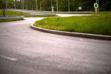 A low-angle view of a curved asphalt ramp on a highway interchange. The road is bordered by a concrete curb and a green grass median with road signs in the background.