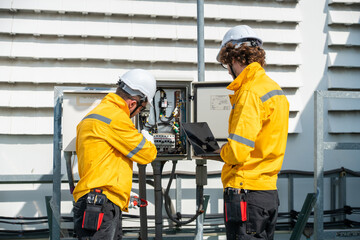 Engineers team in safety helmets and yellow jackets inspecting electrical control cabinet with laptop and tablet on site. Mobile phone Telecommunication Radio antenna tower installed on building