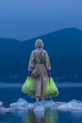 Fototapeta premium A woman is walking on the water, wearing an oversized beige trench coat and purple gloves, holding green bags in her hand with plastic trash inside. 