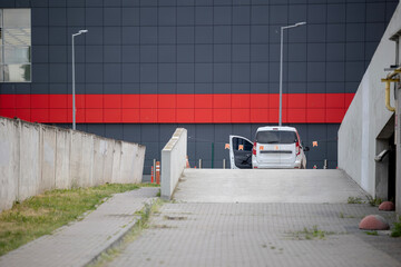 A white van is parked at the top of a concrete ramp leading to an underground garage. In the background is the modern facade of a large building with grey and red panels.