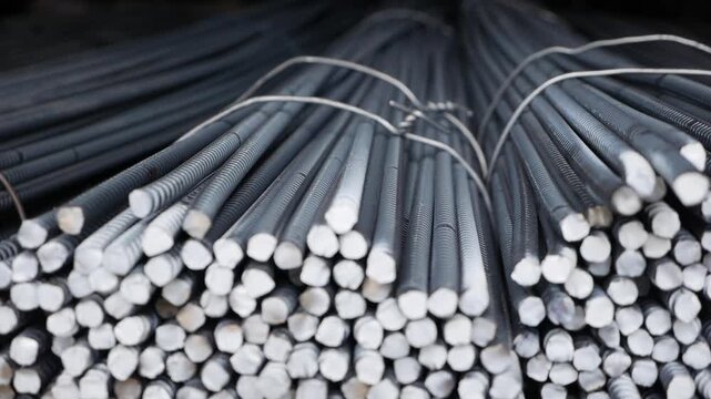 Steel rods stacked neatly in a warehouse. The rods are cylindrical, metallic, and have a shiny surface. They are bundled together with wire ties.