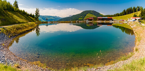 High resolution stitched alpine summer panorama with reflections in a lake and a religious cross at...