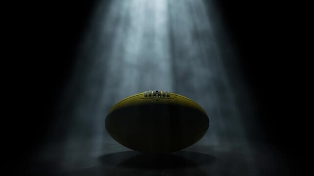 A generic aussie rules sports ball being lit by a single rotating and swirling ethereal light ray on a dark black background