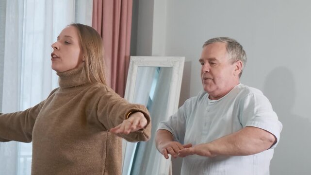Adult daughter teaching her elderly father practicing slow balance exercises indoors with arms stretched out. Wellness at home, family support, senior fitness, healthy lifestyle, active seniors