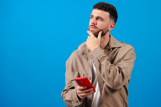 Thoughtful young man in beige jacket holds red smartphone against vivid blue studio background,hand on beard, gazing upward while brainstorming and drafting new social media message on the app today
