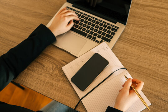 Person working at wooden desk with laptop, writing in a notebook with golden pen, smartphone placed on notebook, modern office environment