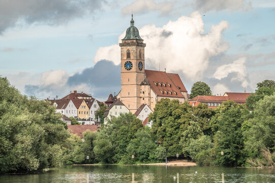 H&auml;userzeile mit N&uuml;rtinger Stadtkirche St. Laurentius am Neckarufer mit Sandstrand und gr&uuml;nen Laubb&auml;umen vor Wolkengebilde an hellblauem Himmel 