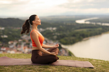 Side view of woman doing yoga in beautiful mountain location with city and river view, sitting cross-legged while meditating, pilates workout in mountains for wellness