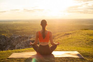 Fototapeta premium Back view of woman meditating in yoga asana in the mountains at the sunset outdoors. Spiritual practice concept