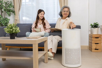 Asian elderly woman and young woman sitting on sofa in bright living room, happily using modern air purifier to improve indoor air quality