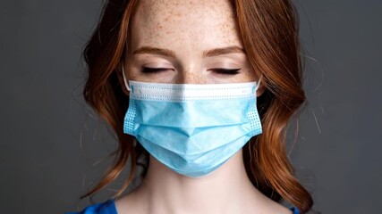 A close-up depicts a woman with striking red hair, freckles, and blue eyes, wearing a medical mask against a gray backdrop