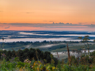 Obraz premium The sun rises over a dreamy, misty landscape filled with rolling hills and trees.