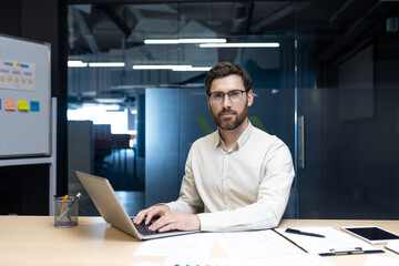 Portrait of a young man working in an office, sitting at a desk with a laptop and looking intently at the camera