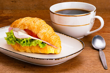 Croissant ham cheese in white ceramic tray with dark edge, metal fork and a cup of black coffee on wooden table.