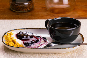 Side view of a cup of coffee in black ceramic cup with blueberry cheese pie on foil paper and metal spoon in white ceramic tray on wooden table.
