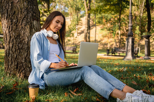 Young woman student working studying while sitting on grass in city park using laptop wireless headphones writing notes. Female freelancer telecommuting, chatting online and e-learning outdoors - Powered by Adobe