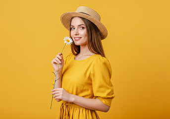 A beautiful smiling woman in a yellow summer dress and straw hat, enjoying the sun and smelling a daisy flower.