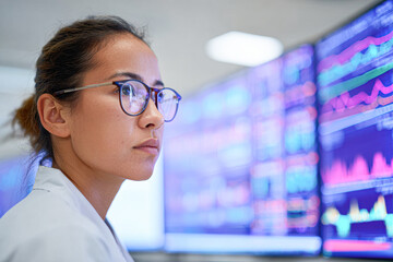 A woman in a lab coat monitors data on multiple digital screens displaying colorful graphs and charts in a high-tech environment.