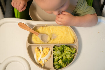 Top-down shot shows curious toddler in feeding chair picking nutritious foods - corn cereal, steamed veggies and egg. Important early eating skills development.