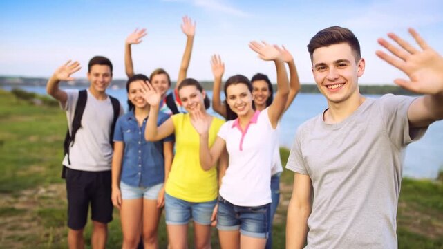 A group of young adults with outstretched hands waves towards the viewer outdoors, smiles, summer day