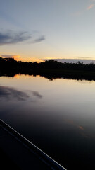 Obraz premium Cuiabá River in Pantanal wetlands at dusk with orange sunset reflecting on the calm water surface. A dark silhouette of the forest on the horizon under a twilight sky. Peaceful natural landscape view.