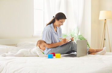 Fototapeta premium Cheerful smiling happy young mother in casual working on a laptop computer while her cute little child playing with toys lying on bed in bedroom at home. Motherhood and education concept.