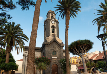 The Iglesia de Nuestra Se&ntilde;ora de la Pe&ntilde;a de Francia in Puerto de la Cruz, Tenerife, framed by tall palm trees and a clear blue sky, showcasing its iconic clock tower and historic stone facade.