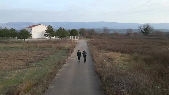 Una pareja camina con su perro por una carretera tranquila mientras el dron los sigue desde atr&aacute;s.